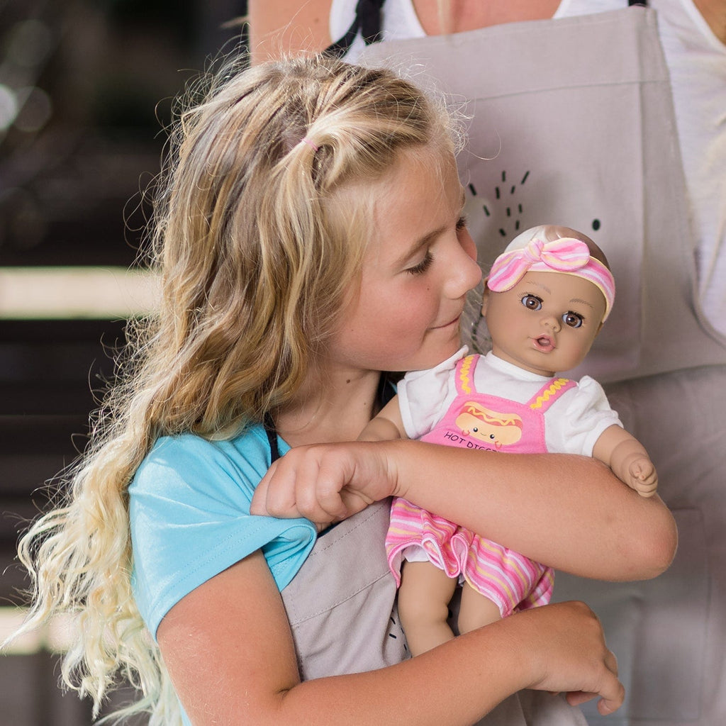 Adora Hot Diggity Dog baby doll set includes one 13-inch doll, a hot dog-printed one-piece dress with white undershirt, matching headband, white bloomers, and a pink baby bottle. Made for Ages 1+.